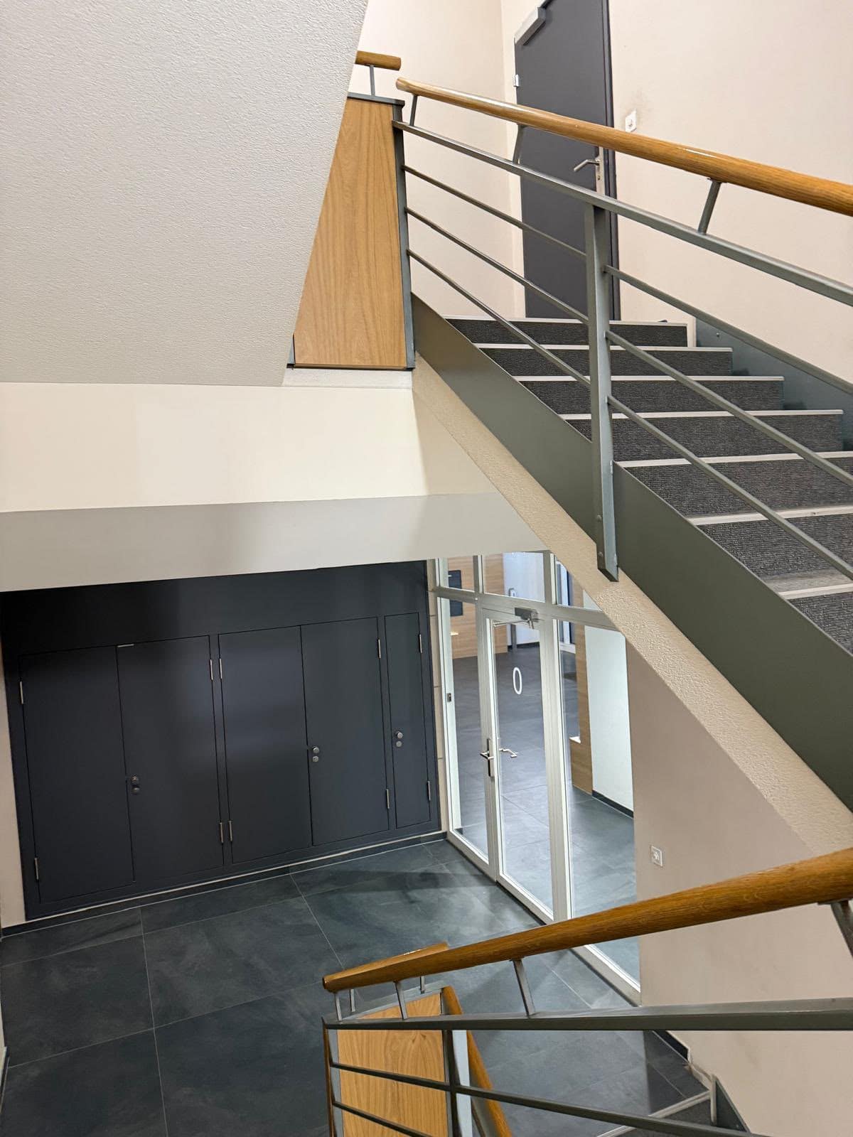 Treppenhaus mit Stahlgeländer und Handlauf aus Holz, unten eine Glastür zur Eingangshalle und dunkler Plattenboden
