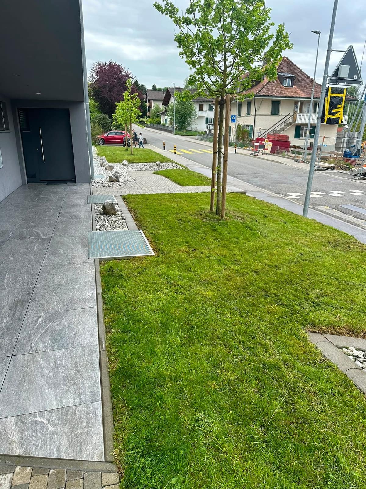 Eingangsbereich einer Neubauliegenschaft mit frisch gemähter Rasenfläche, Kiesstreifen und Lichtschächten