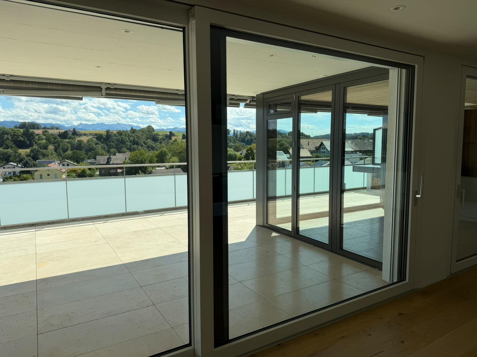 Raumhohe Schiebefenster und eine verglaste Ecke zu einer Dachterrasse mit Glasgeländer, dahinter Hügel und Berge