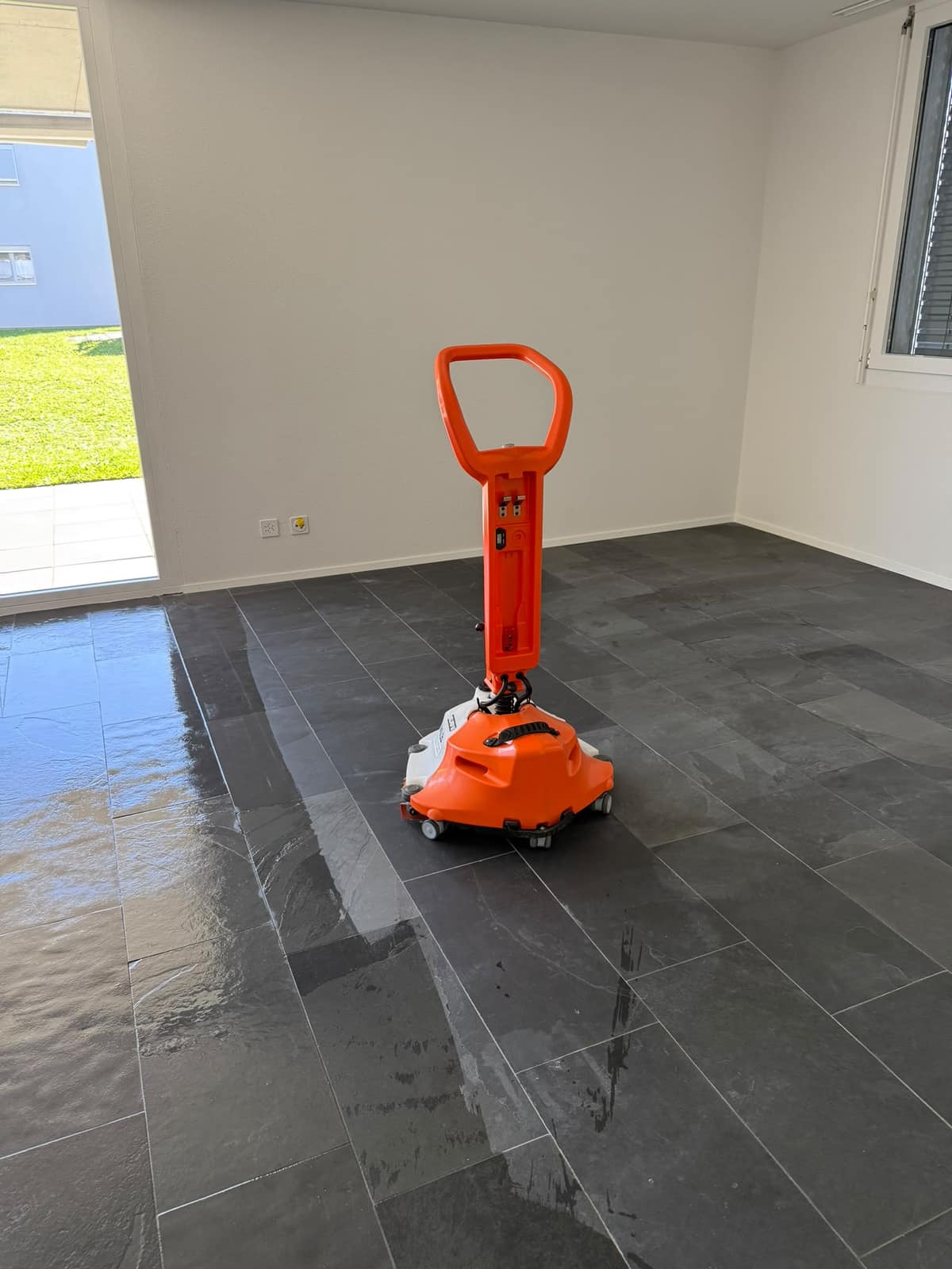 Orange Reinigungsmaschine auf einem nassen, dunklen Plattenboden in einem leeren Zimmer